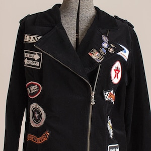 Op de afbeelding: Zwarte corduroy jas versierd met diverse patches, waaronder "MY WAY HIGHWAY", "FORD MOTOR Co." en "MUSTANG". De jas heeft een ritssluiting en meerdere decoratieve spelden. Een Route 66-patch is ook zichtbaar.