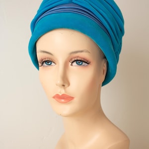 Puede incluir: Un turbante turquesa con un dise&ntilde;o plisado y una banda de sat&eacute;n. El sombrero se exhibe en una cabeza de maniqu&iacute; con ojos azules y labios carnosos. El sombrero es de estilo vintage.