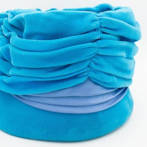 Puede incluir: Un turbante azul turquesa con un dise&ntilde;o fruncido y drapeado. El sombrero presenta una banda de tela azul claro alrededor de la base. La tela parece ser de terciopelo suave.