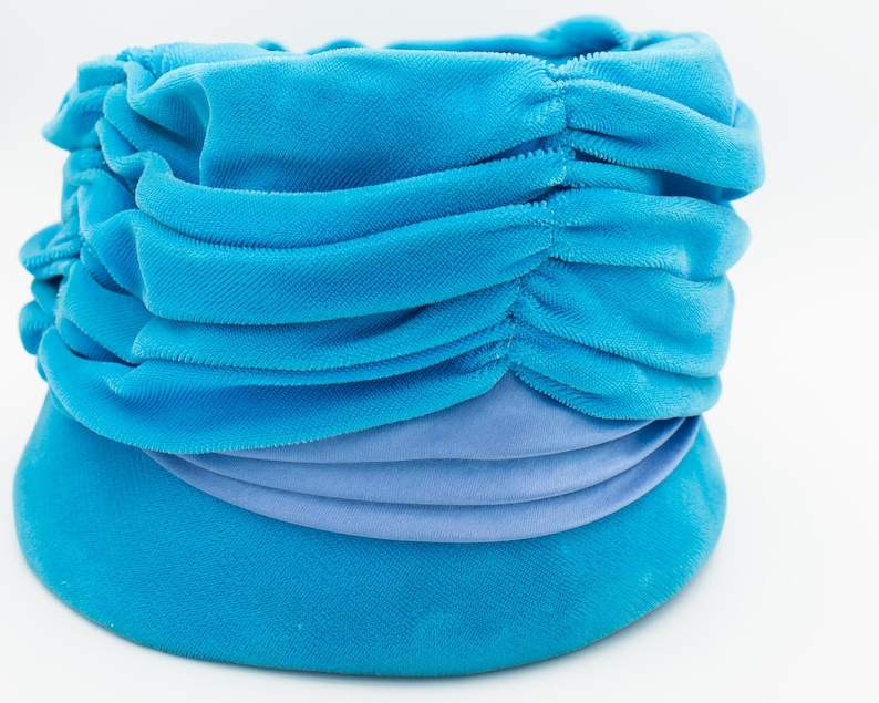Puede incluir: Un turbante azul turquesa con un dise&ntilde;o fruncido y drapeado. El sombrero presenta una banda de tela azul claro alrededor de la base. La tela parece ser de terciopelo suave.