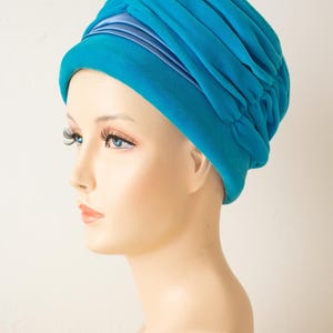 Puede incluir: Un turbante turquesa con un dise&ntilde;o fruncido. El sombrero presenta una banda de tela azul claro alrededor de la base y se muestra en una cabeza de maniqu&iacute;. El sombrero est&aacute; hecho de un material suave y fluido.