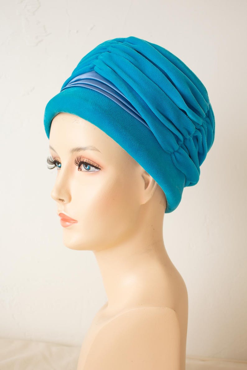 Puede incluir: Un turbante turquesa con un dise&ntilde;o fruncido. El sombrero presenta una banda de tela azul claro alrededor de la base y se muestra en una cabeza de maniqu&iacute;. El sombrero est&aacute; hecho de un material suave y fluido.
