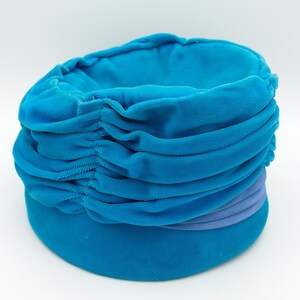 Puede incluir: Un sombrero estilo turbante azul turquesa con un dise&ntilde;o de tela fruncida. El sombrero presenta una banda de tela morada clara alrededor del centro. El sombrero est&aacute; hecho de un material suave y aterciopelado.