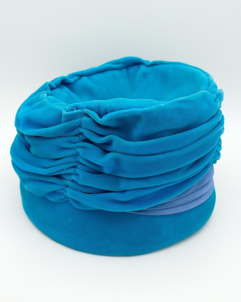 Puede incluir: Un sombrero estilo turbante azul turquesa con un dise&ntilde;o de tela fruncida. El sombrero presenta una banda de tela morada clara alrededor del centro. El sombrero est&aacute; hecho de un material suave y aterciopelado.