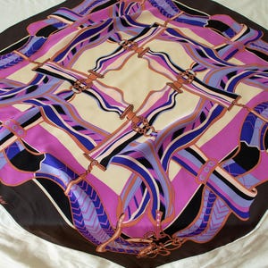 Puede incluir: Pañuelo cuadrado de seda con un estampado geométrico en tonos rosa, morado, azul, negro y beige. El pañuelo presenta un diseño repetitivo de formas y líneas estilizadas, con un borde marrón oscuro y elementos decorativos.