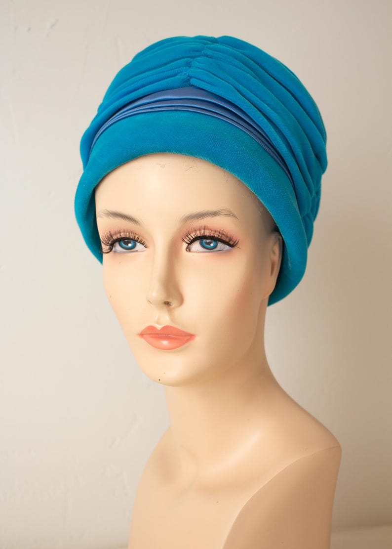 Puede incluir: Un turbante turquesa con un dise&ntilde;o plisado y una banda de sat&eacute;n. El sombrero se exhibe en una cabeza de maniqu&iacute; con ojos azules y labios carnosos. El sombrero es de estilo vintage.