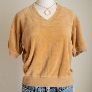 Puede incluir: Una camiseta de color mostaza con cuello en V y mangas cortas, hecha de un material suave y aterciopelado. La camiseta se combina con vaqueros azules. Un collar dorado con un colgante circular se lleva con la camiseta. La camiseta se muestra en un maniquí.