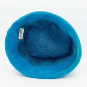 Puede incluir: Un sombrero turquesa vintage con una corona redondeada y un interior azul pato oscuro. El sombrero presenta un fino forro de malla y un ribete de cinta verde azulado. Una etiqueta con texto es visible dentro del sombrero. El sombrero est&aacute; hecho de un material suave y aterciopelado.