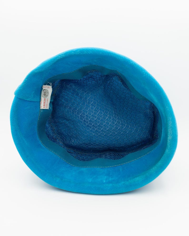 Puede incluir: Un sombrero turquesa vintage con una corona redondeada y un interior azul pato oscuro. El sombrero presenta un fino forro de malla y un ribete de cinta verde azulado. Una etiqueta con texto es visible dentro del sombrero. El sombrero est&aacute; hecho de un material suave y aterciopelado.