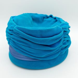 Puede incluir: Un sombrero turquesa estilo turbante con una corona redondeada y un ala ancha y plana. El sombrero presenta tela fruncida y una banda de tela azul m&aacute;s clara alrededor de la base. El sombrero est&aacute; hecho de un material suave y aterciopelado.