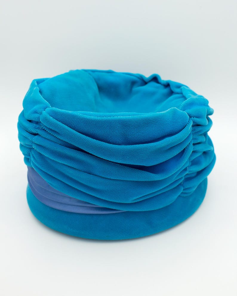 Puede incluir: Un sombrero turquesa estilo turbante con una corona redondeada y un ala ancha y plana. El sombrero presenta tela fruncida y una banda de tela azul m&aacute;s clara alrededor de la base. El sombrero est&aacute; hecho de un material suave y aterciopelado.