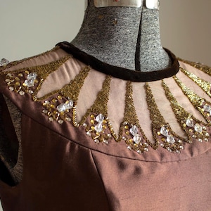 Könnte beinhalten: Ein braunes Kleidoberteil mit verziertem Ausschnitt. Der Ausschnitt hat einen Samtbesatz, goldene Stickereien und klare Kristallakzente. Das Kleid wird auf einer Schaufensterpuppe präsentiert.