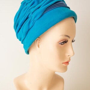 Puede incluir: Un sombrero estilo turbante azul turquesa con un dise&ntilde;o fruncido. El sombrero presenta una banda azul oscuro alrededor de la base. El sombrero se exhibe en una cabeza de maniqu&iacute;.