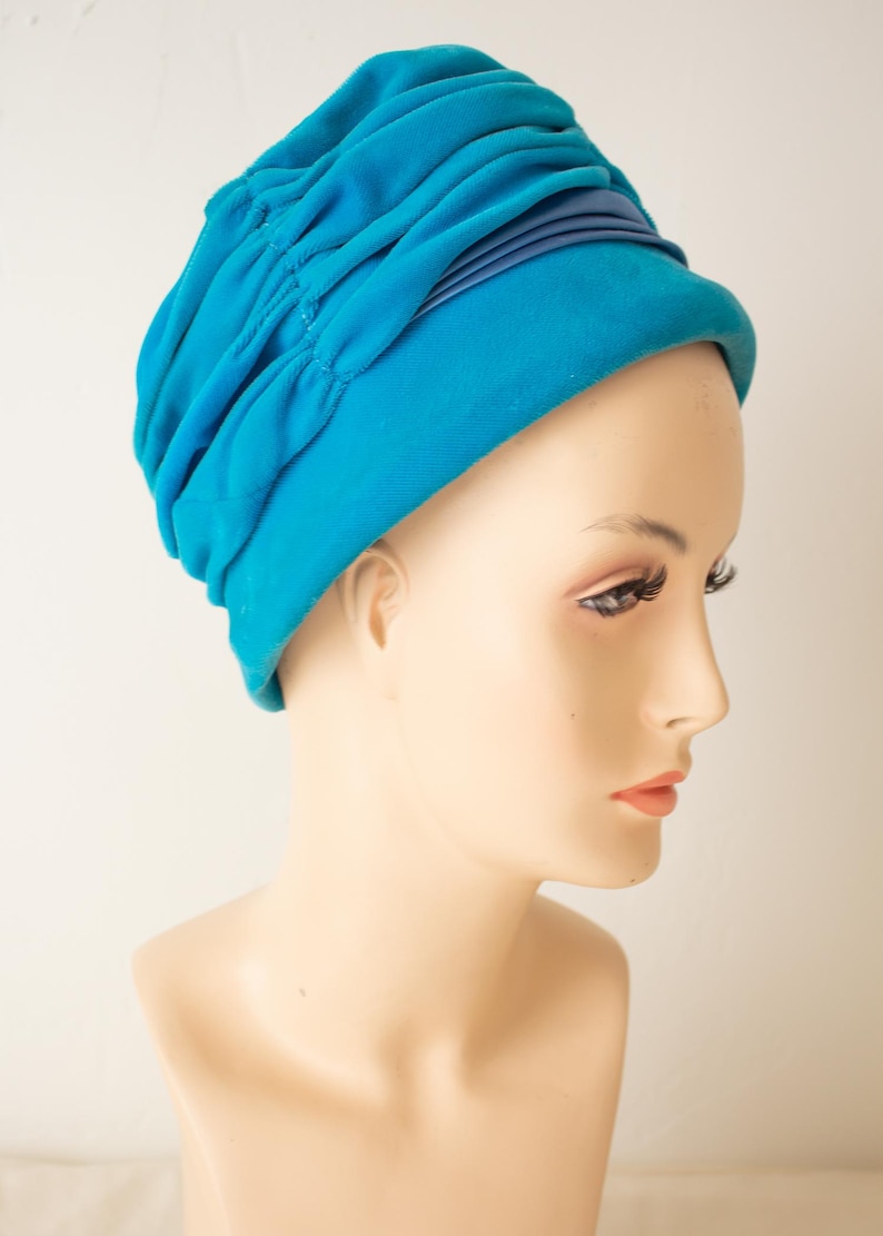 Puede incluir: Un sombrero estilo turbante azul turquesa con un dise&ntilde;o fruncido. El sombrero presenta una banda azul oscuro alrededor de la base. El sombrero se exhibe en una cabeza de maniqu&iacute;.