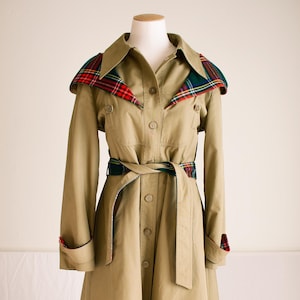 May include: A tan trench coat with a plaid collar and cuffs. The coat features a button-up front, a belted waist, and long sleeves. The plaid pattern includes red, green, and yellow accents. The coat is displayed on a mannequin.