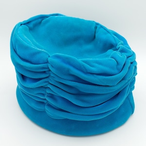 Puede incluir: Un vibrante sombrero tipo turbante turquesa. El sombrero presenta una corona redondeada con detalles horizontales de tela fruncida. El ala es una banda lisa y s&oacute;lida. El sombrero est&aacute; hecho de un material suave y aterciopelado, sobre fondo blanco.