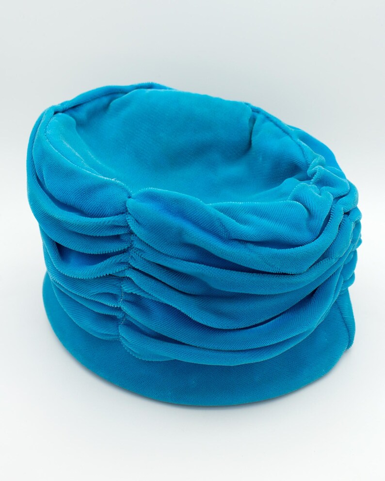 Puede incluir: Un vibrante sombrero tipo turbante turquesa. El sombrero presenta una corona redondeada con detalles horizontales de tela fruncida. El ala es una banda lisa y s&oacute;lida. El sombrero est&aacute; hecho de un material suave y aterciopelado, sobre fondo blanco.