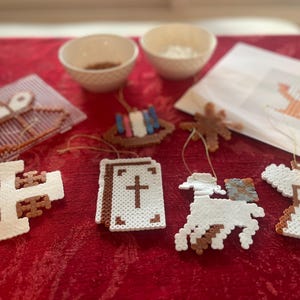 May include: A collection of colorful beaded ornaments with religious themes, including a cross, a bible, an angel, and a lamb. The ornaments are made with white, brown, and blue beads and are strung on white thread.
