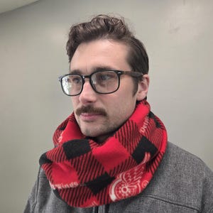May include: Red and black plaid infinity scarf. The scarf features a buffalo plaid pattern with a white paisley design and the word "Detroit" printed on it. The scarf is made of a soft, warm material.