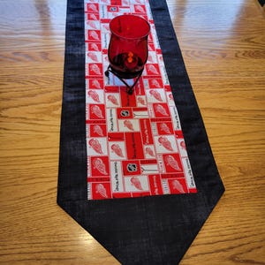 May include: A black and red table runner with a repeated pattern of the Detroit Red Wings logo. The runner is on a wooden table with a red glass candle holder in the center.