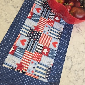 May include: A patriotic table runner with a patchwork design featuring red, white, and blue stars, stripes, and hearts. The runner has a navy blue border with white star accents. A red bowl filled with fruit sits on the table.