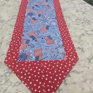 May include: A patriotic table runner with a red border featuring white stars. The center has a light blue background with American flags and fireworks. This decorative runner is perfect for Independence Day.