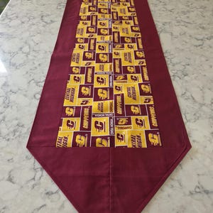 May include: A maroon and gold table runner featuring a repeating pattern of gold squares with the text "CENTRAL MICHIGAN CHIPPEWAS" and a "C" logo. The runner has a maroon border and a pointed end, suitable for a dining table or decorative display.