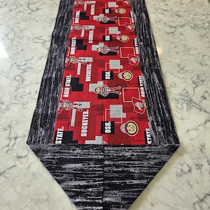May include: A decorative table runner featuring a red and black pattern with the text "OHIO STATE" and "BUCKEYES". The center design includes OSU logos and figures. The runner is approximately 60 inches long.