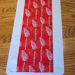 May include: Red and white table runner with a repeating pattern of the Detroit Red Wings logo. The runner has a white border and is made of fabric.