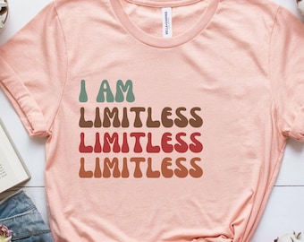 May include: A pale pink t-shirt featuring the text "I AM LIMITLESS" in a stacked design. The words are in teal, brown, red, and orange. The shirt is made of a soft material.