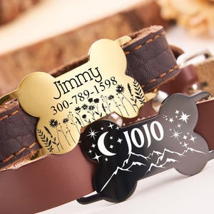 May include: Two personalized dog tags on brown leather collars. The gold tag reads "Jimmy 300-789-1598" with a floral design. The black tag reads "Jojo" with a crescent moon and mountain design.