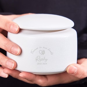 Può includere: Un'urna bianca in ceramica per animali domestici con coperchio, tenuta in mano. L'urna è incisa con le parole "Forever in our hearts", un'impronta di zampa e un simbolo di cuore, il nome "Ripley" e gli anni "2016-2024."