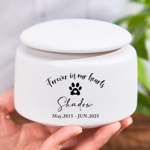 Könnte beinhalten: Weiße Keramik-Urne für Haustiere mit Deckel. Die Urne trägt den Text "Forever in our hearts" über einem Pfotenabdruck und den Namen "Shadow" mit den Daten May.2015 - JUN.2025.