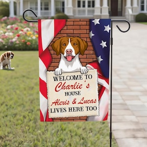 May include: A garden flag featuring a dog illustration with an American flag design. The flag reads "Welcome to Charlie's House" with additional names. A real dog sits in the background.
