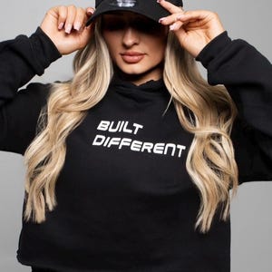 May include: A black cropped hoodie with the text "BUILT DIFFERENT" in white. The model is wearing a black mesh baseball cap with a white "B" logo and a black mini skirt.