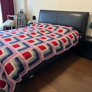 Może przedstawiać: Czarne skórzane łóżko z różową, czerwoną i niebieską dzianą narzutą. Łóżko znajduje się w sypialni z drewniana podłogą i czarną szafką nocną.