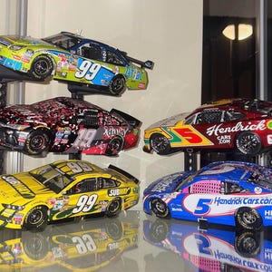 Può includere: Una collezione di cinque modelli di auto da corsa NASCAR in vari colori, tra cui giallo, rosso e blu. Ogni auto presenta loghi di sponsor e numeri di gara. Le auto sono esposte su una mensola.