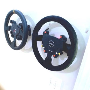May include: Two black racing steering wheels mounted on a white wall. One wheel has a gold accent and the other has the Sparco logo. Both have multiple buttons and controls.