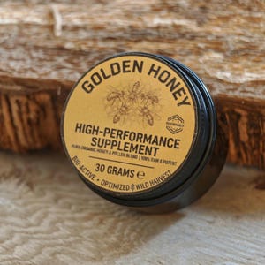 Może przedstawiać: Czarny słoik ze złotą etykietą z napisem "GOLDEN HONEY HIGH-PERFORMANCE SUPPLEMENT". Etykieta zawiera również ilustrację pszczół i napis "30 GRAMÓW". Słoik stoi na drewnianej powierzchni.
