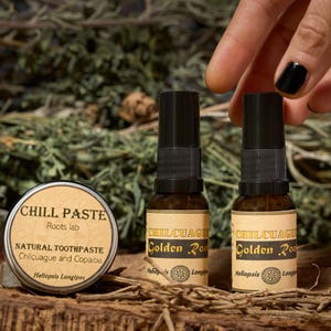 Op de afbeelding: Een afbeelding met natuurlijke gezondheidsproducten. Een ronde blik van "CHILL PASTE" natuurlijke tandpasta en twee kleine flesjes "Golden Root" extract worden getoond. De producten staan op een houten oppervlak met groen op de achtergrond.