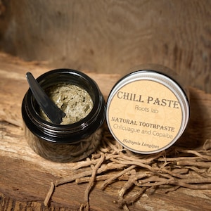 Può includere: Due piccoli vasetti neri di dentifricio naturale, uno aperto con un applicatore nero. L'etichetta recita "CHILL PASTE" e "NATURAL TOOTHPASTE Chilcuague and Copaiba". I vasetti sono su una superficie di legno con radici essiccate.