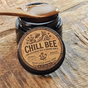 Puede incluir: Un tarro de miel cruda de raíz de chilcuague y miel orgánica Chill Bee con una cuchara de madera encima. La tapa del tarro está quitada, revelando la miel. La etiqueta incluye texto sobre ingredientes y tamaño de la porción. El tarro está sobre una superficie de madera.