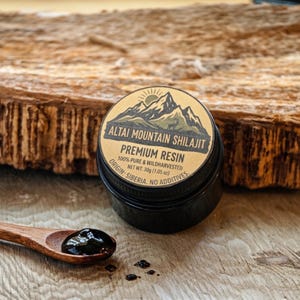Könnte beinhalten: Ein kleiner, schwarzer Tiegel mit Altai Mountain Shilajit Premium Resin, mit einem Etikett, das eine Bergillustration zeigt. Ein Holzlöffel hält einen Teil des dunklen Harzes, mit ein paar Tropfen in der Nähe. Auf dem Etikett steht "100% Pure & Wildharvested, Netto-Gewicht 30g".