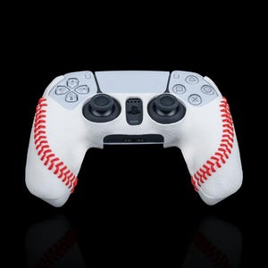 Puede incluir: Un mando de videojuegos blanco y gris con un diseño de béisbol. El mando presenta costuras rojas a los lados, imitando una pelota de béisbol. Los botones y joysticks son negros, sobre un fondo negro.