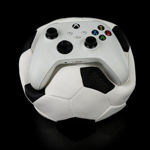 May include: A white Xbox controller rests on top of a soccer ball. The controller has black joysticks and colored buttons. The soccer ball is white with black pentagons.
