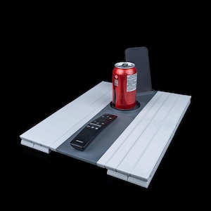 Puede incluir: Una bandeja blanca y gris con un portavasos y una ranura para teléfono integrados. Una lata de refresco roja, un mando a distancia negro y un smartphone están en la bandeja. La bandeja está diseñada para guardar objetos.