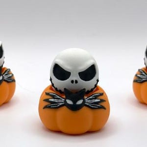Könnte beinhalten: Drei kleine Figuren mit einem weißen, schädelartigen Kopf mit schwarzen Augen und einem genähten Mund, die in einer leuchtend orangefarbenen Kürbisbasis sitzen. Jede Figur enthält ein schwarzes, fledermausartiges Detail mit weißen Akzenten vorne.