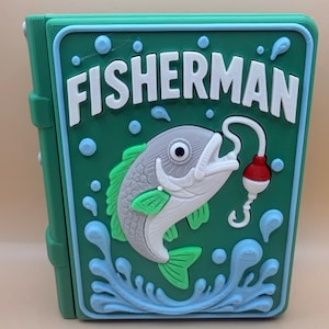Puede incluir: Una caja verde con la palabra "FISHERMAN" en letras blancas. La caja presenta una imagen 3D de un pez con un anzuelo y un flotador. El pez es gris, blanco y verde, con detalles de agua azules.
