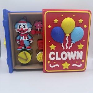 Könnte beinhalten: Ein farbenfrohes Clown-Spielzeug-Set in einer Holzkiste. Der Deckel ist rot mit gelben Akzenten und trägt das Wort "CLOWN" sowie Ballon-Illustrationen. Das Set enthält eine Clownfigur, eine Trommel, eine Blume und weiteres Zubehör.