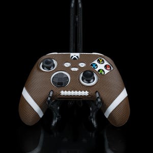 May include: A brown and white football-themed video game controller with a textured surface. The controller has white stripes and a white stitched design. The buttons are colored and the controller is on a black stand.